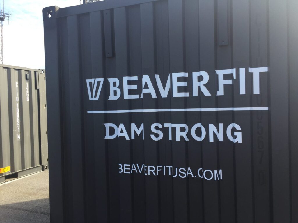 White BeaverFit lettering, Dam Strong slogan and beaverfitusa.com web address applied across the ribs of a black shipping container.