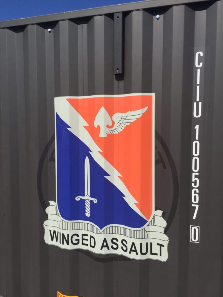 Close-up of the Winged Assault crest graphic, with a sword, lightning bolt and winged arrowhead, conforming to the corrugations of a black steel container.