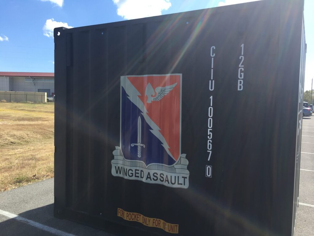 Red, white and blue Winged Assault crest graphic installed on the side of a black corrugated shipping container beside its container number.