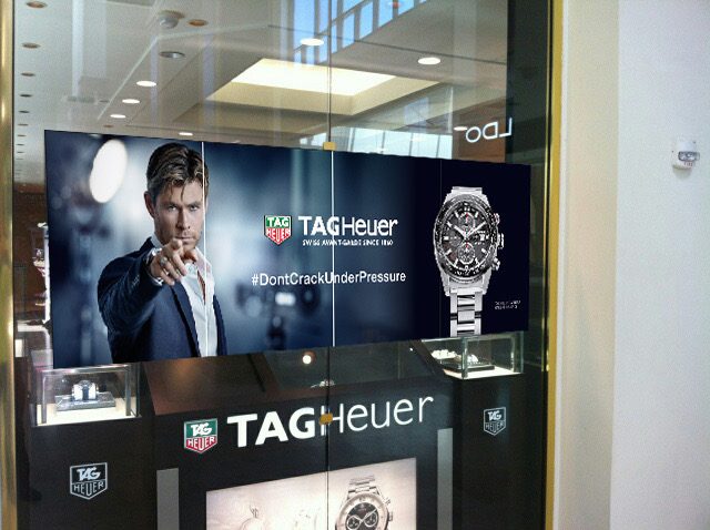 TAG Heuer retail display graphics in a mall store window, with a wide campaign banner above branded watch display cases.
