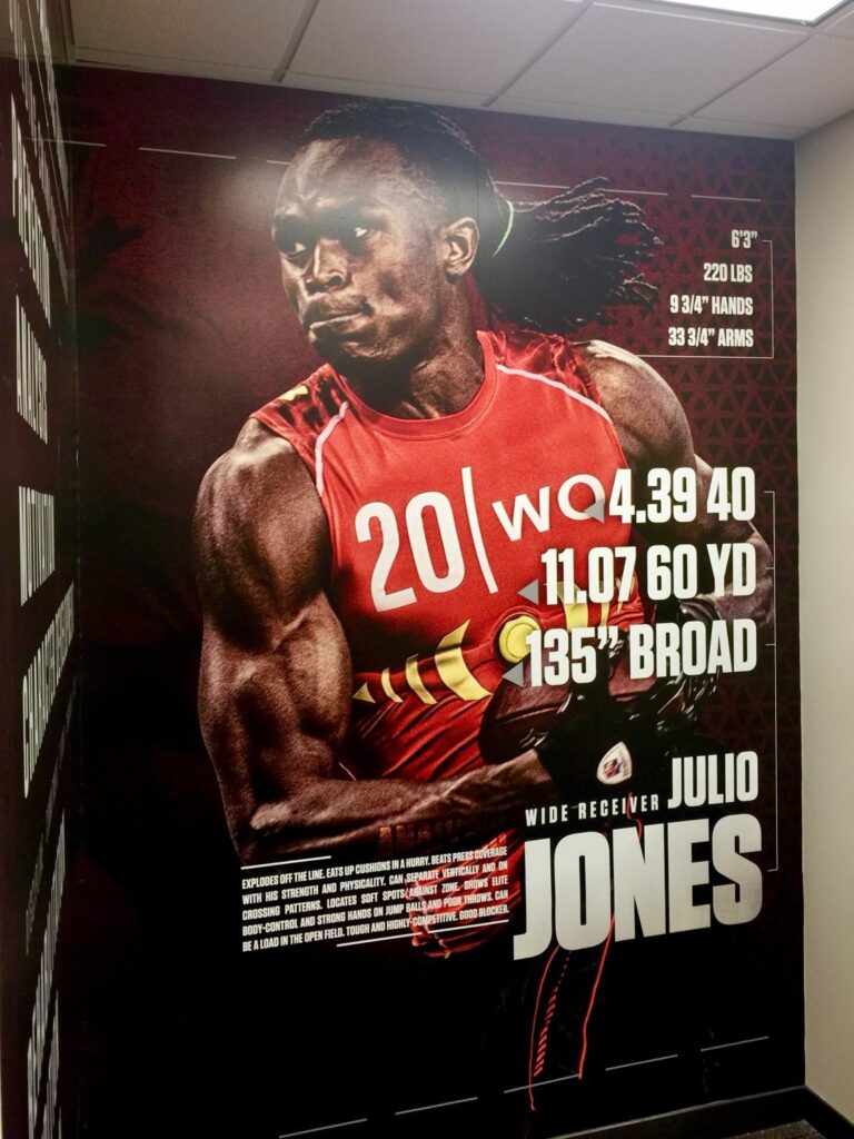 Front view of oversized Julio Jones football mural on a crimson University of Alabama athletic facility wall with printed athlete profile figures