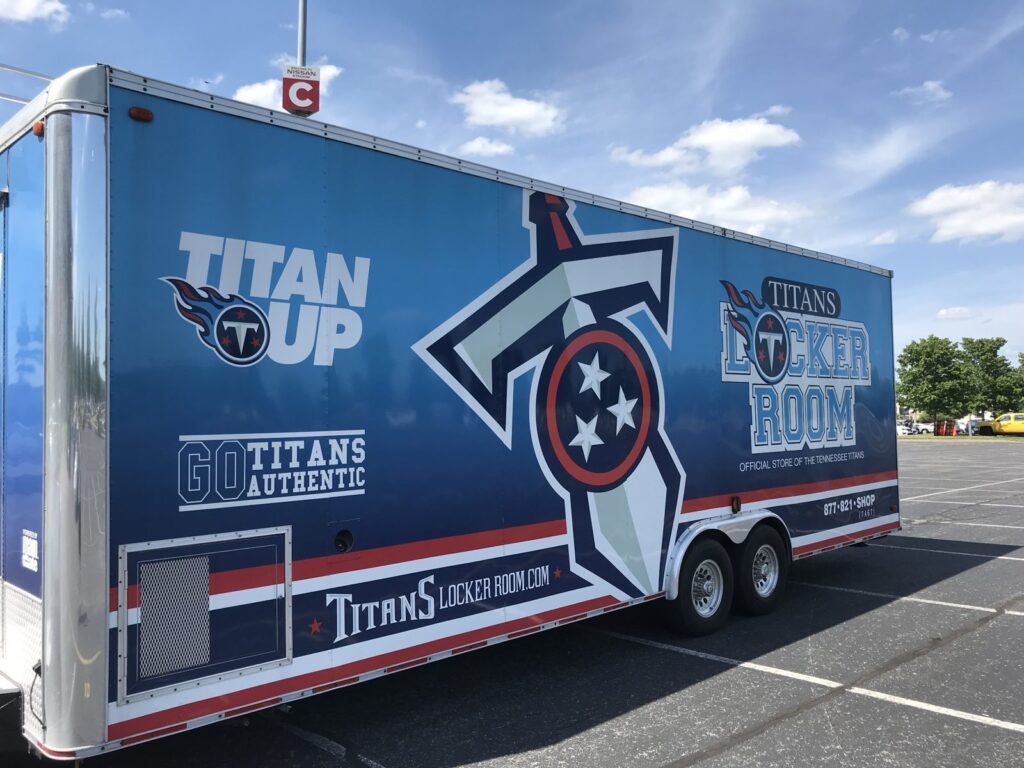 Angled view of the Tennessee Titans trailer wrap showing Titan Up, Go Titans Authentic, TitansLockerRoom.com and the oversized Titans logo across the trailer side in a Nissan Stadium parking lot.