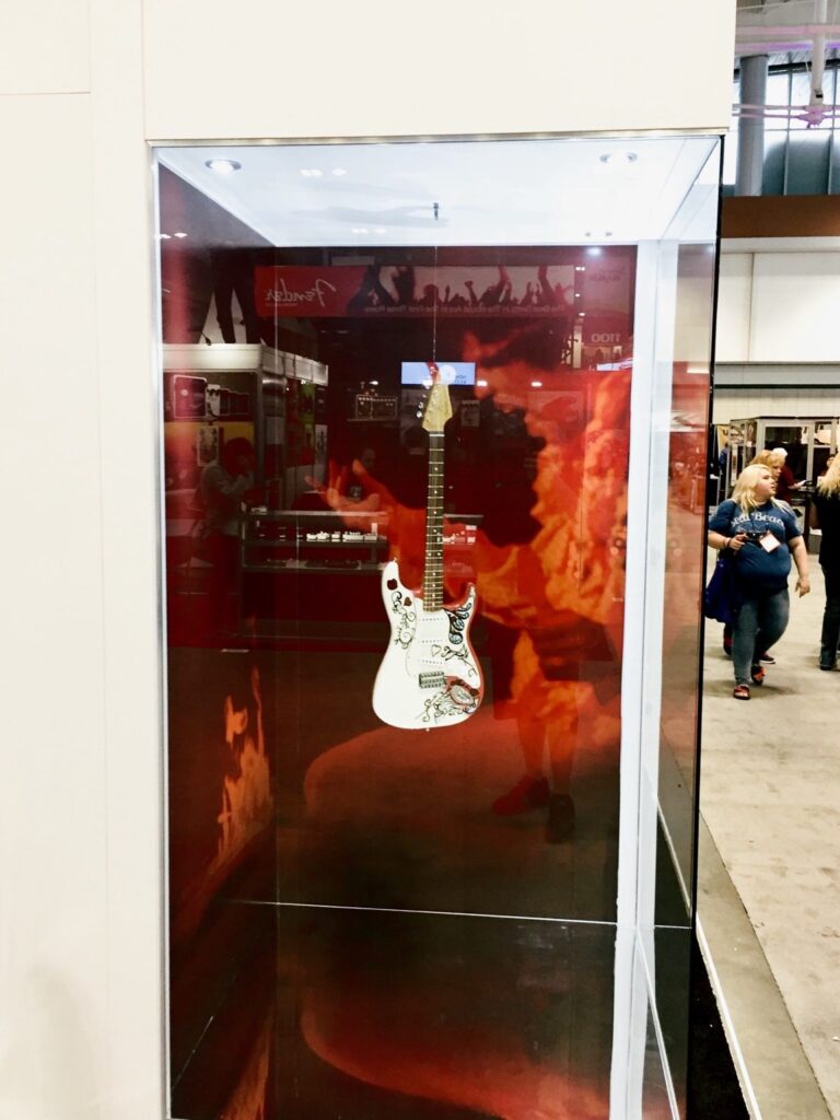 Glass guitar display showcase with red-and-black graphic backdrop inside the Fender booth in Nashville.