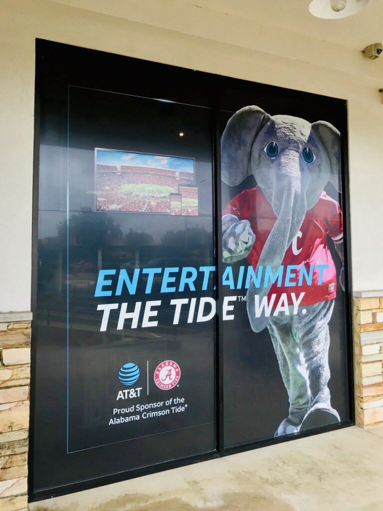 Closer view of AT&T Crimson Tide storefront graphics on two large window panes with elephant mascot, football stadium image, and blue-and-white Entertainment the Tide Way lettering.