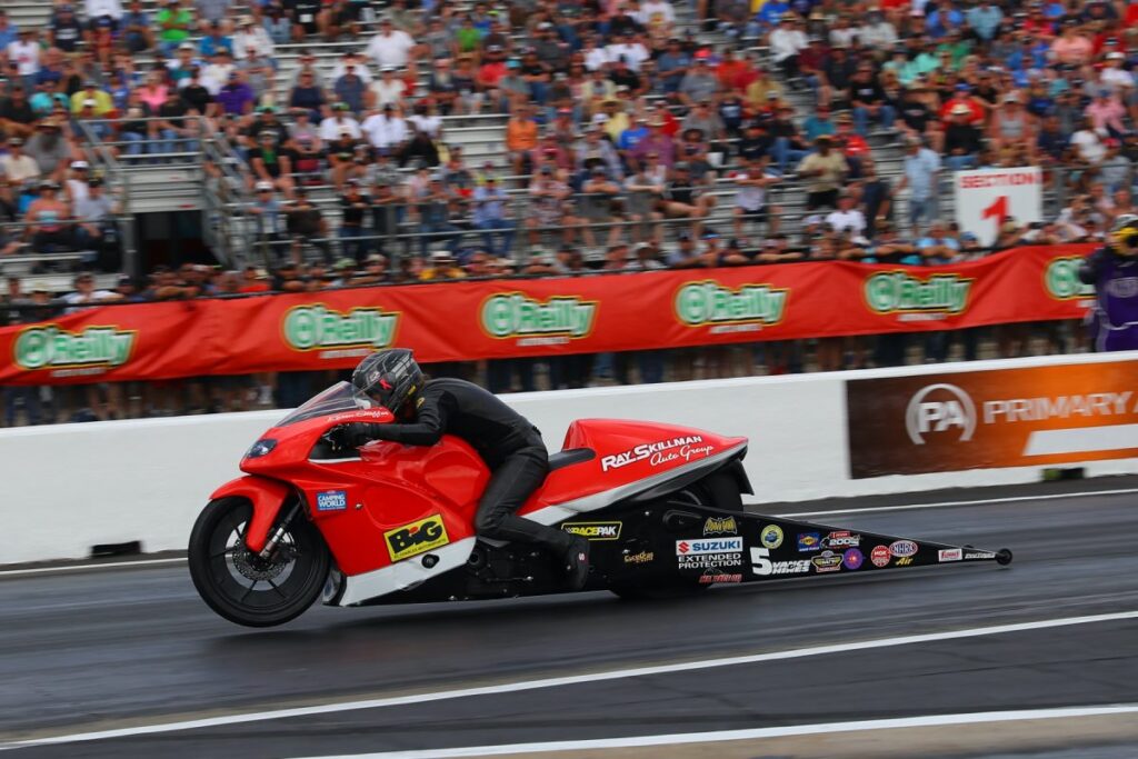 Karen Stoffer racing a red White Alligator Racing Suzuki Pro Stock Motorcycle down a drag strip in front of a full grandstand