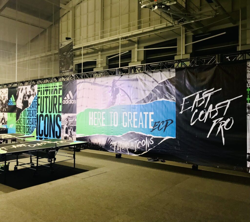 Indoor East Coast Pro backdrop banners with Adidas Here to Create graphics hung on truss beside table tennis tables.