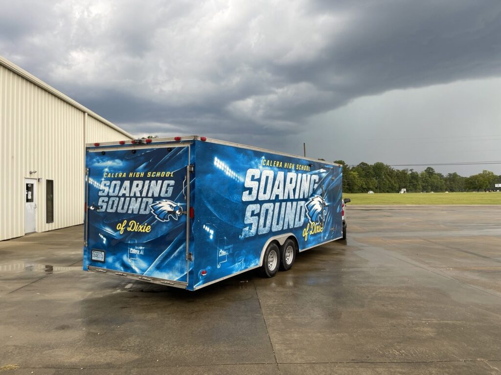Wide rear three-quarter photograph of completed blue Calera High School Soaring Sound of Dixie cargo trailer graphics outside a commercial building.