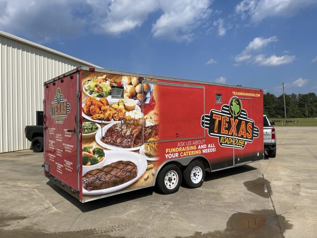 Red Texas Roadhouse enclosed trailer showing rear logo and restaurant locations alongside full-color food imagery and a large side logo.