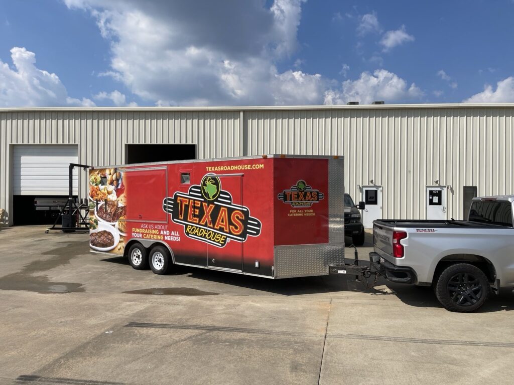 Red Texas Roadhouse cargo trailer hitched to a pickup truck outside a metal-sided commercial workshop in Alabaster, Alabama.