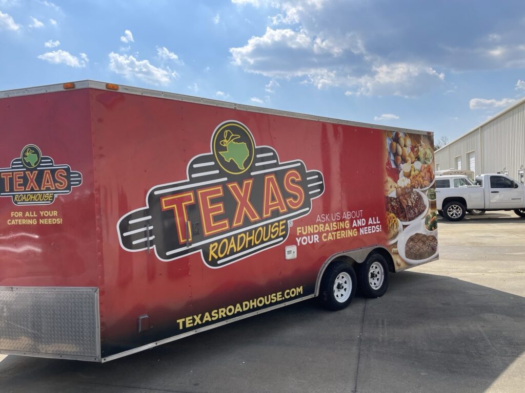 Wide side view of red Texas Roadhouse enclosed cargo trailer with oversized restaurant logo, catering and fundraising graphics, and food photography.