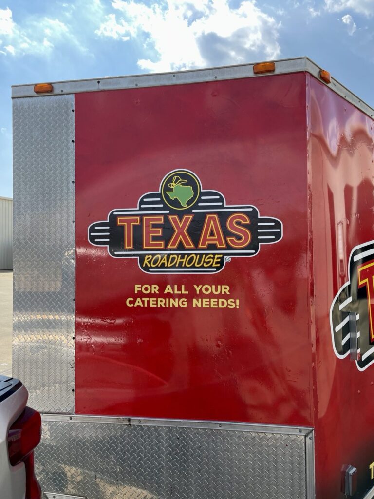 Close-up of the red front panel of a Texas Roadhouse cargo trailer with a restaurant logo, yellow catering message and aluminum diamond-plate corner trim.