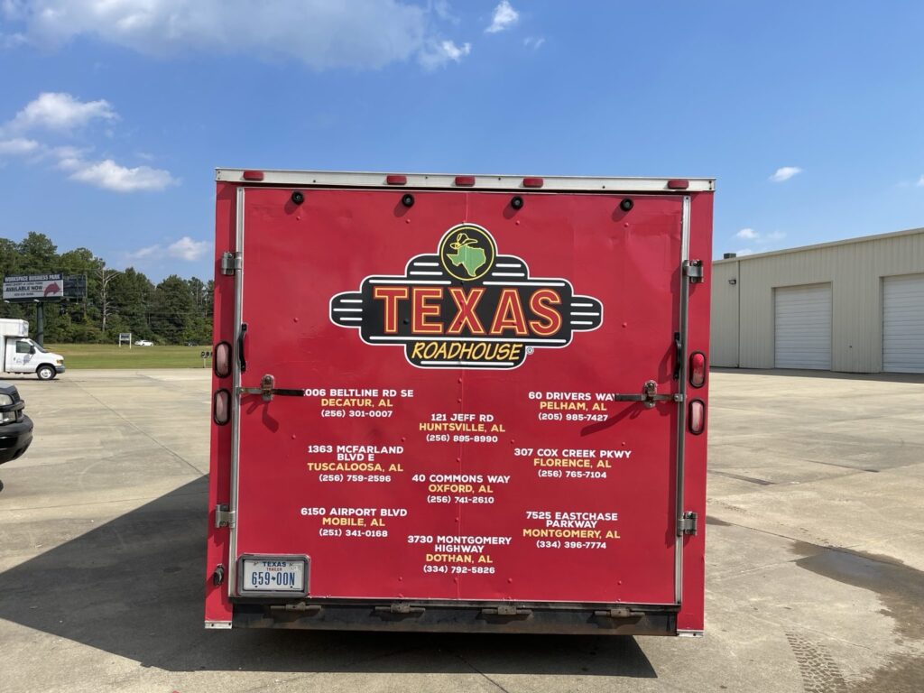 Straight-on view of red Texas Roadhouse enclosed cargo trailer rear doors with restaurant logo and printed contact details for multiple Alabama restaurants, including Pelham.