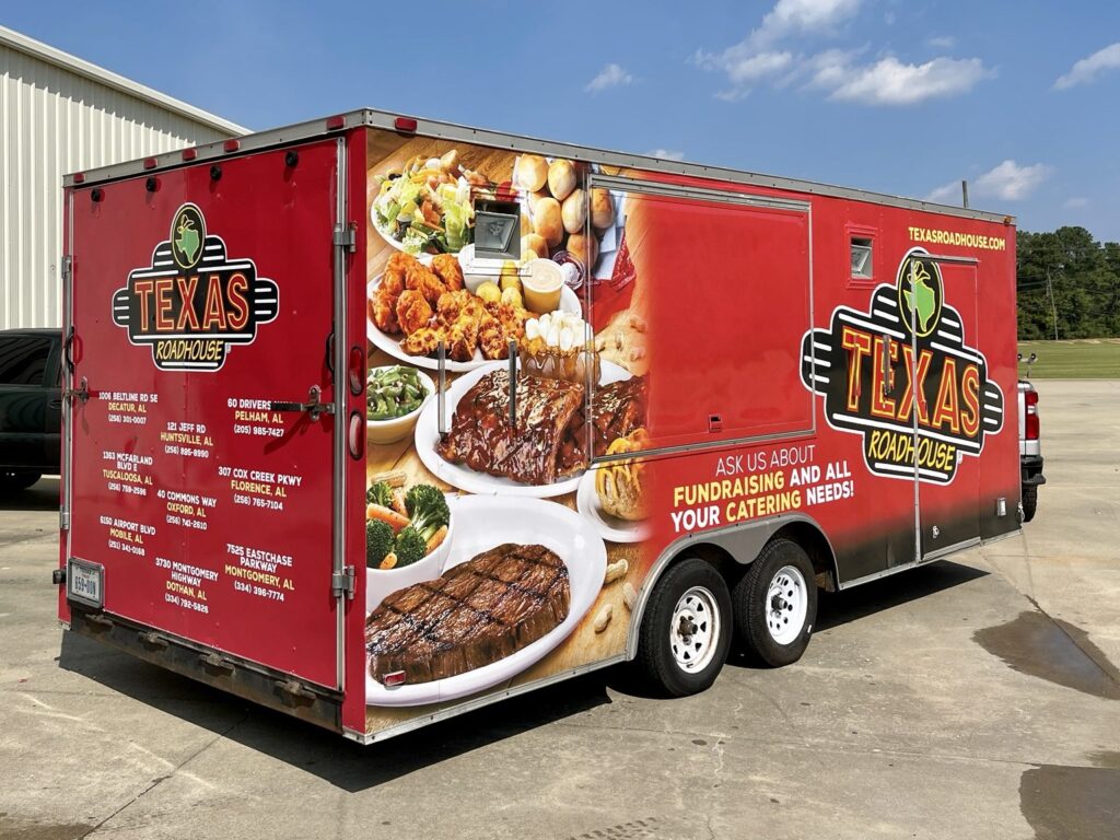 Rear three-quarter view of a red Texas Roadhouse catering trailer with photographs of steak and other dishes on the side and a list of restaurant locations on the rear doors.