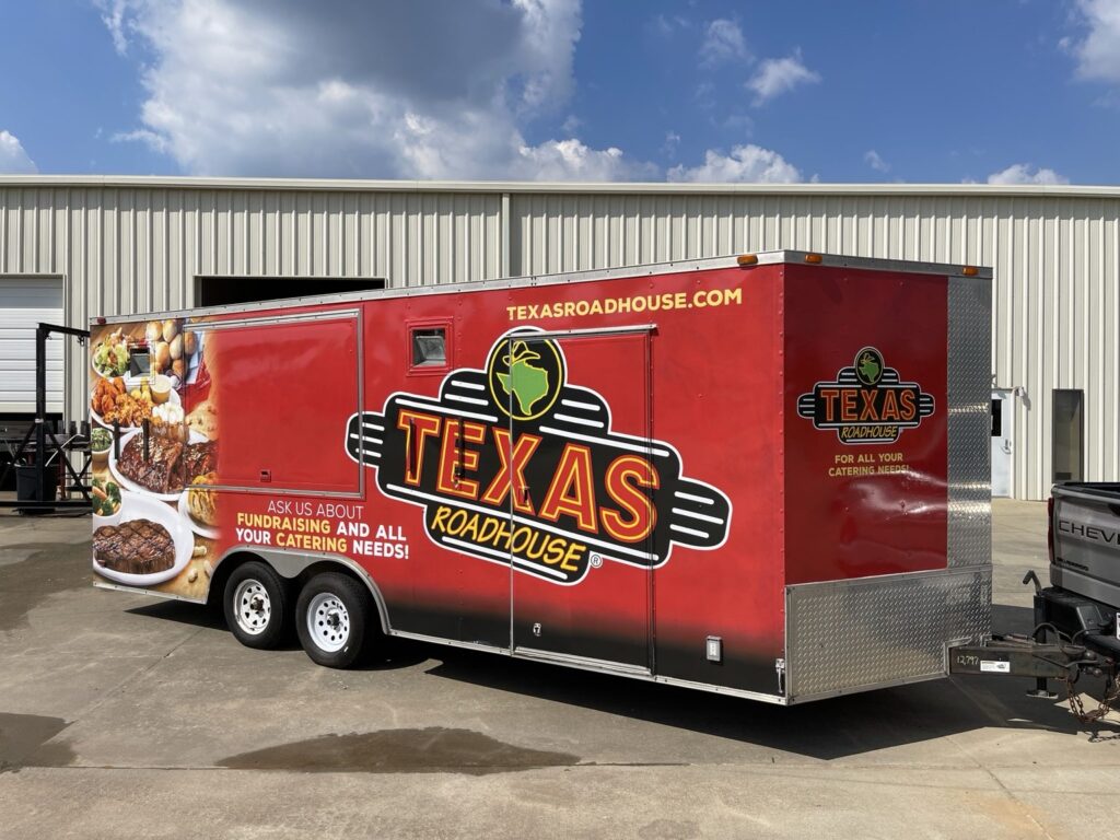 Completed red Texas Roadhouse cargo trailer with large logo graphics positioned across the side access door and service opening, plus restaurant website and food photographs.