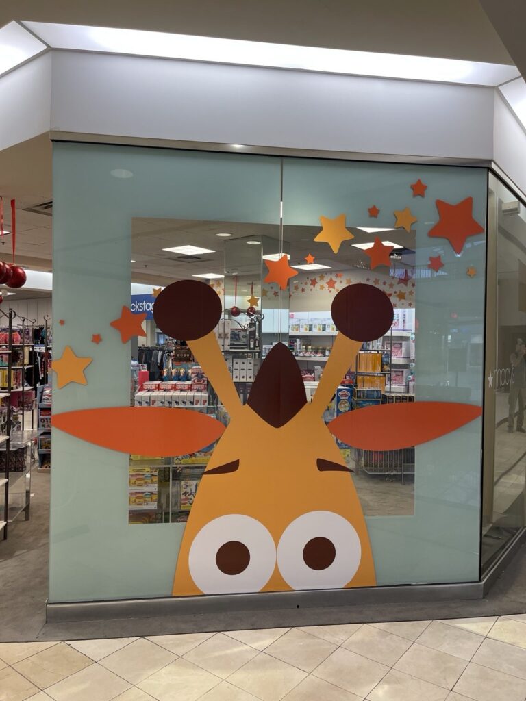 Large orange and yellow Toys R Us giraffe mascot graphic with colorful stars applied across a tall glass panel inside the Macy's store at Riverchase Galleria.