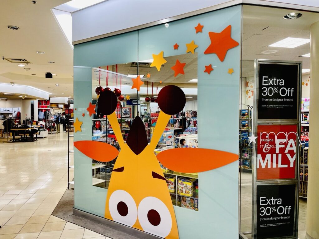 Wide view of Toys R Us giraffe-and-stars window graphics at Macy's in Hoover, Alabama, showing the decorative glass panel and neighboring department store aisles.