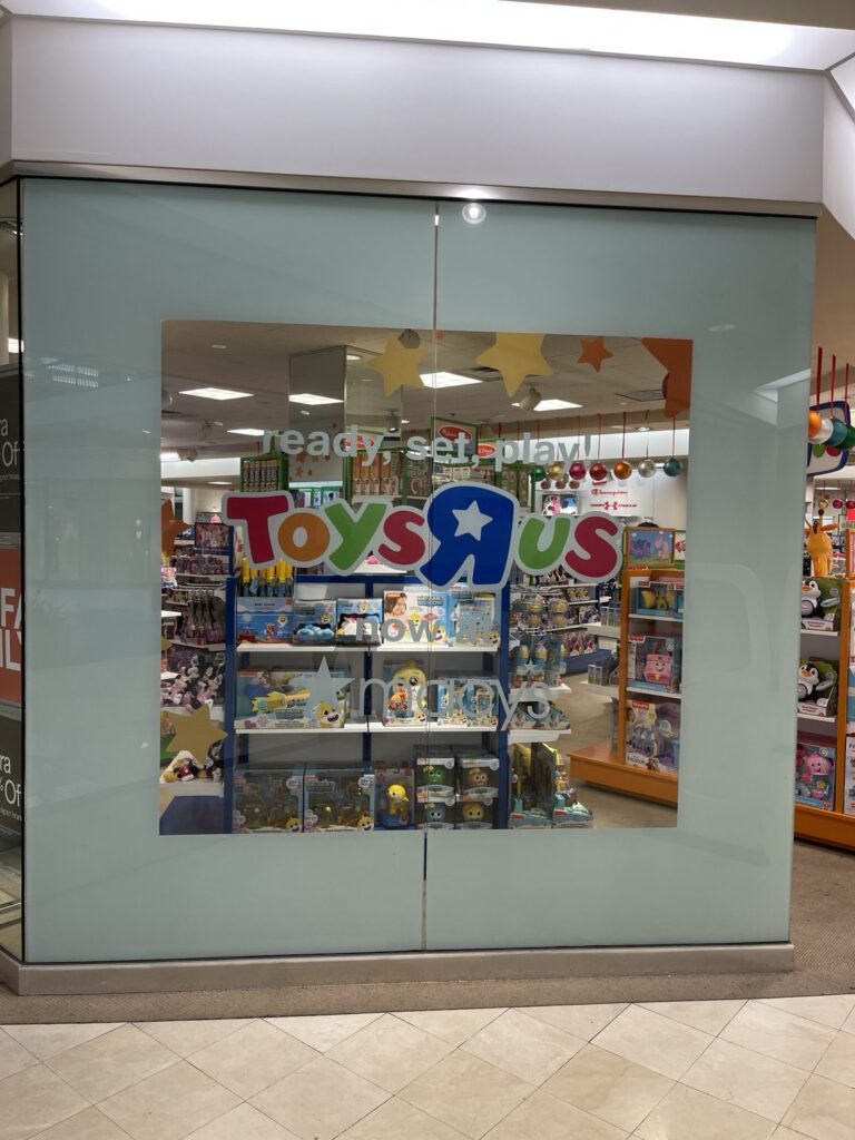 Toys R Us window graphics Hoover: full glass entrance area for Toys R Us inside Macy's, with pale blue window background, centered Toys R Us logo and retail merchandise visible through the graphic opening.