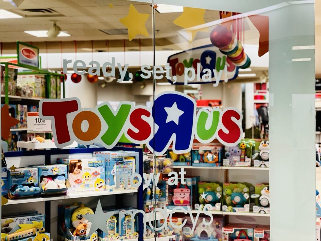 Front angled view of the Toys R Us shop-in-shop at Macy's with colorful window branding, star shapes and now at Macy's message visible on glass.