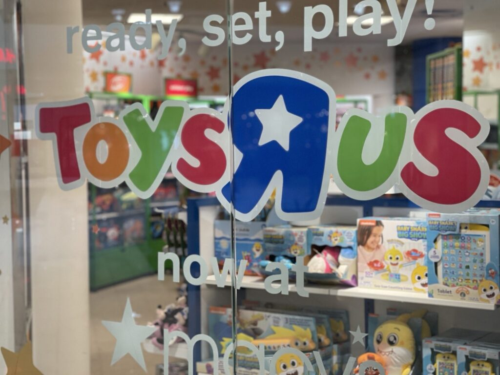 Close-up of colorful Toys R Us wordmark and ready, set, play lettering installed on retail storefront glass with toy displays visible behind.