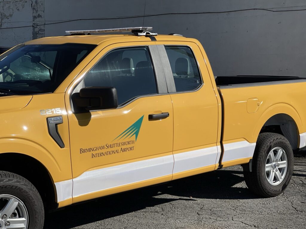 Yellow airport service truck with the airport logo and a white lower-body stripe