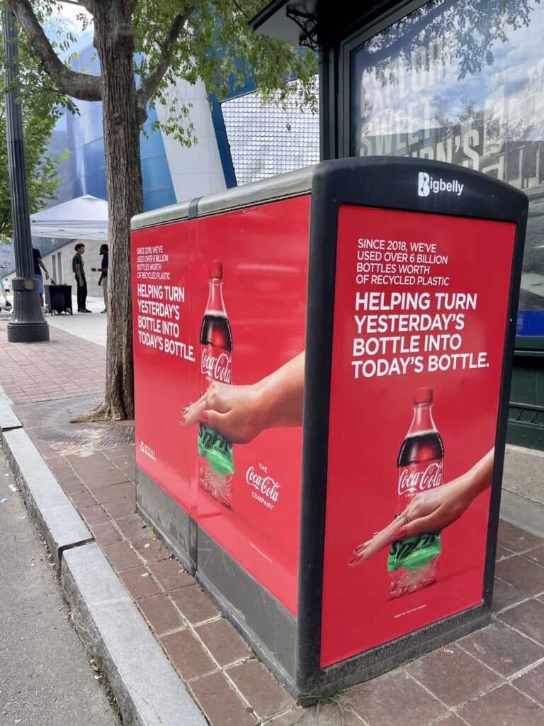 Coca-Cola red sustainability campaign graphics displayed across a black Bigbelly bin along a brick sidewalk in downtown Atlanta.