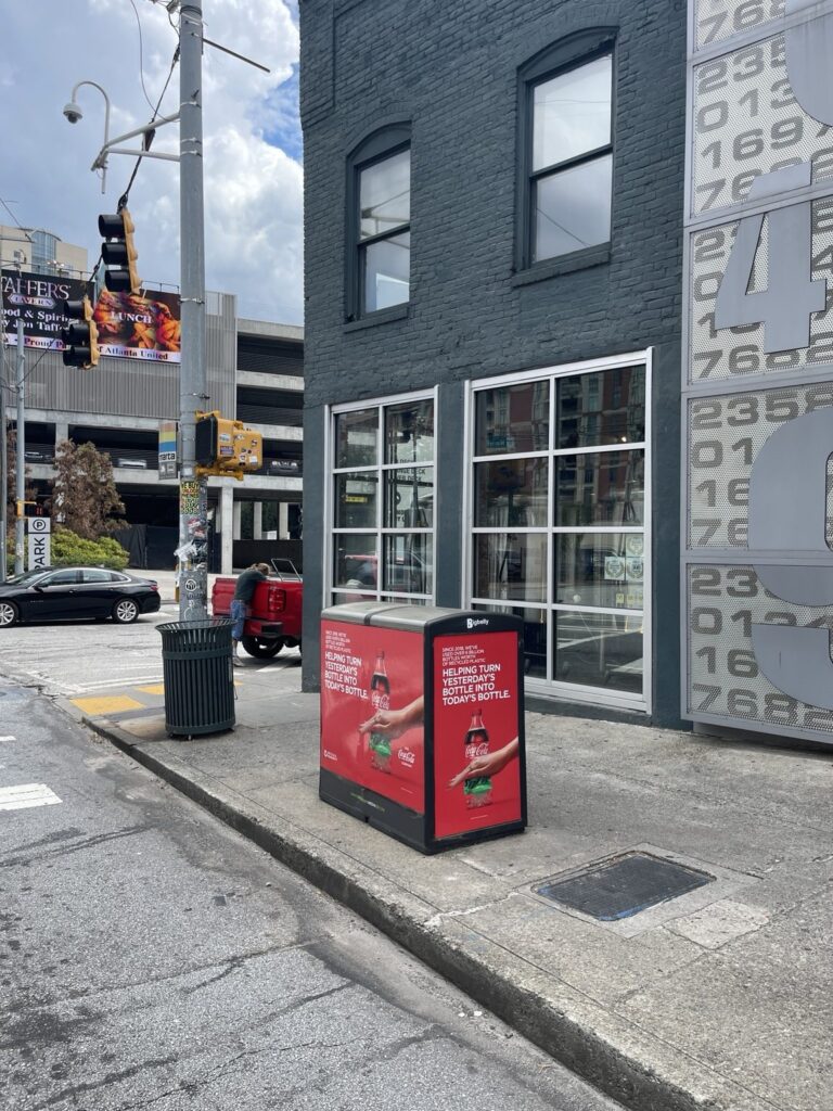 Black Bigbelly street-side bin with red Coca-Cola bottle recycling campaign artwork along a downtown Atlanta sidewalk near a crosswalk.