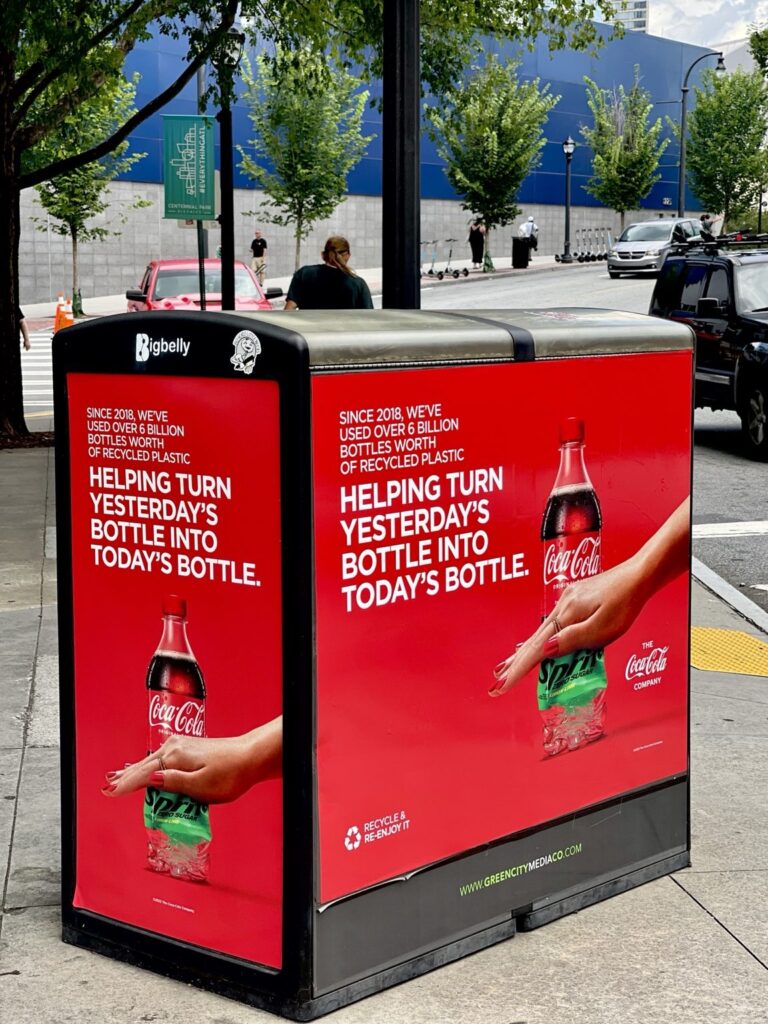 Red Coca-Cola recycled-bottle advertising graphics on multiple visible sides of a black sidewalk Bigbelly bin in downtown Atlanta.