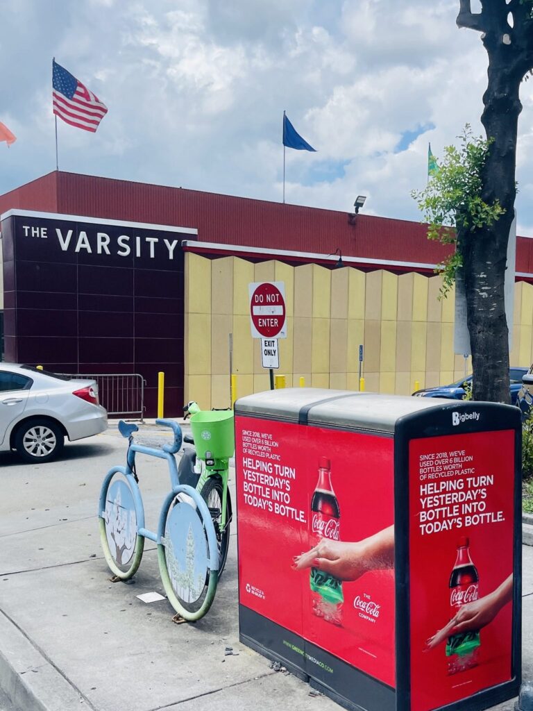 Red Coca-Cola recycled-plastic campaign graphics installed on a black Bigbelly sidewalk waste and recycling bin near The Varsity in Atlanta, Georgia.