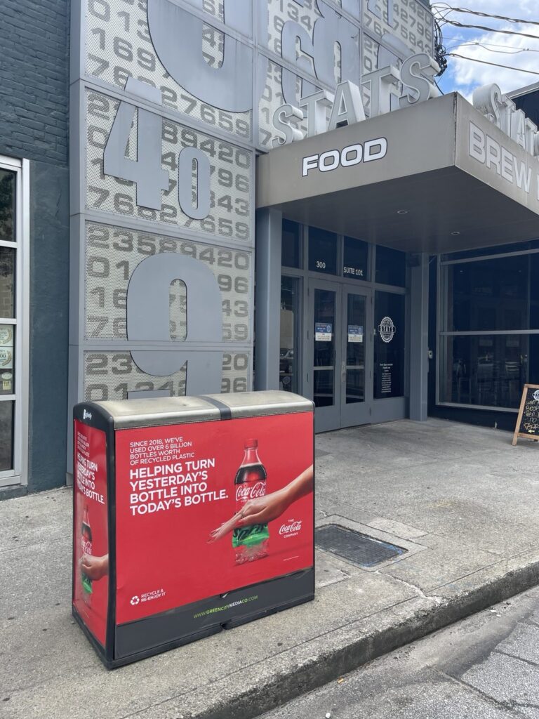 Red Coca-Cola recycled-plastic campaign graphic fitted to a sidewalk waste bin near the STATS restaurant entrance in Atlanta, Georgia.