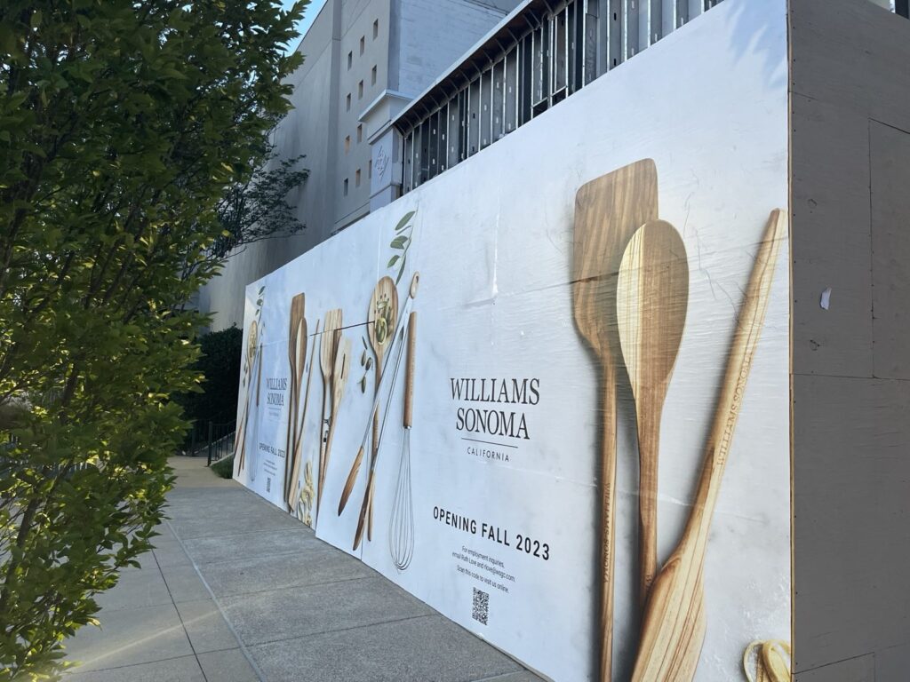 Long perspective of the Williams Sonoma printed retail barricade at The Summit in Birmingham, running beside trees and a pedestrian walkway.