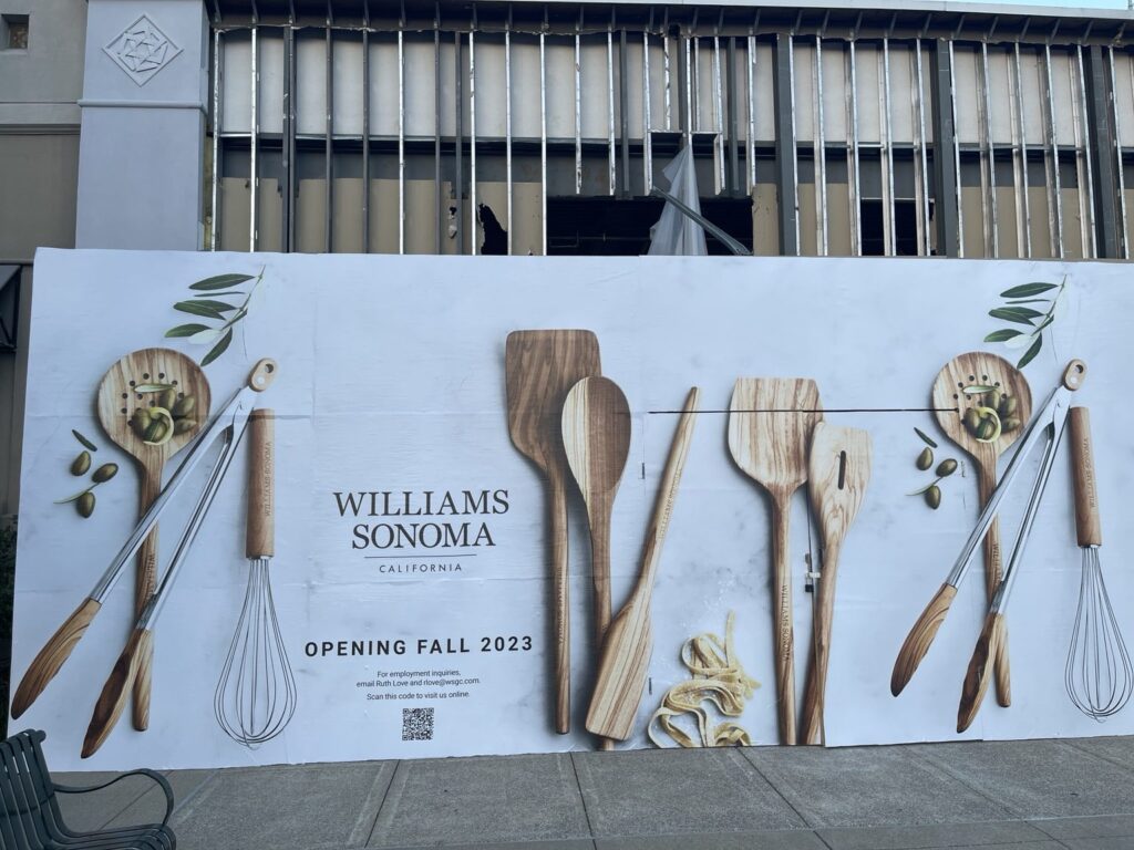 Straight-on view of the Williams Sonoma construction barricade at The Summit with kitchen utensil graphics and exposed storefront renovation framing above.