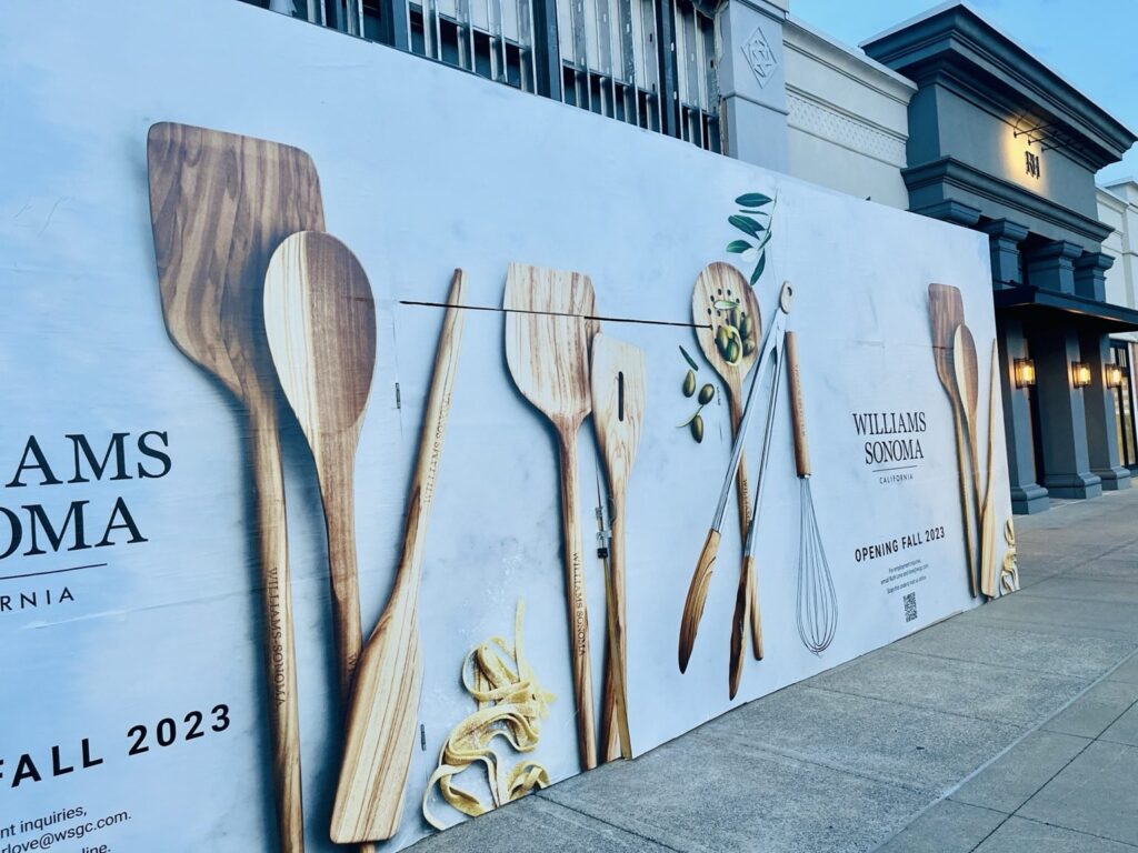 Angled view of a long Williams Sonoma construction barricade wrap at The Summit in Birmingham showing oversized cooking utensils and repeated brand messaging.