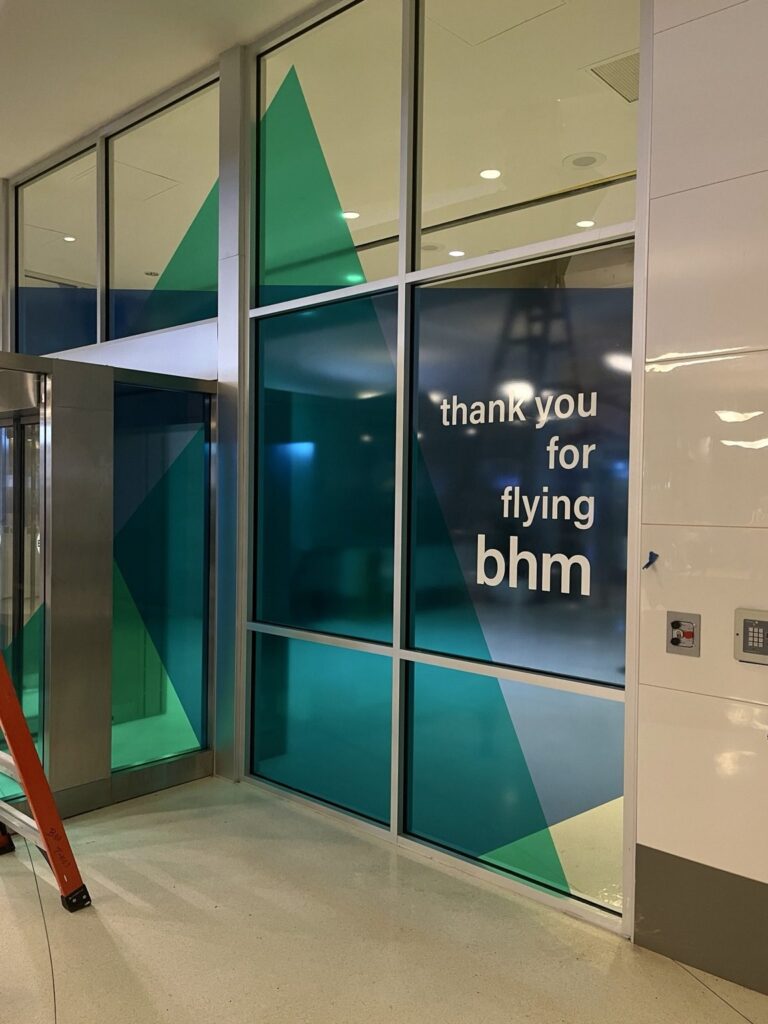 Angled view of the Birmingham airport interior glass wall with layered green and blue geometric film and a white passenger thank-you message.