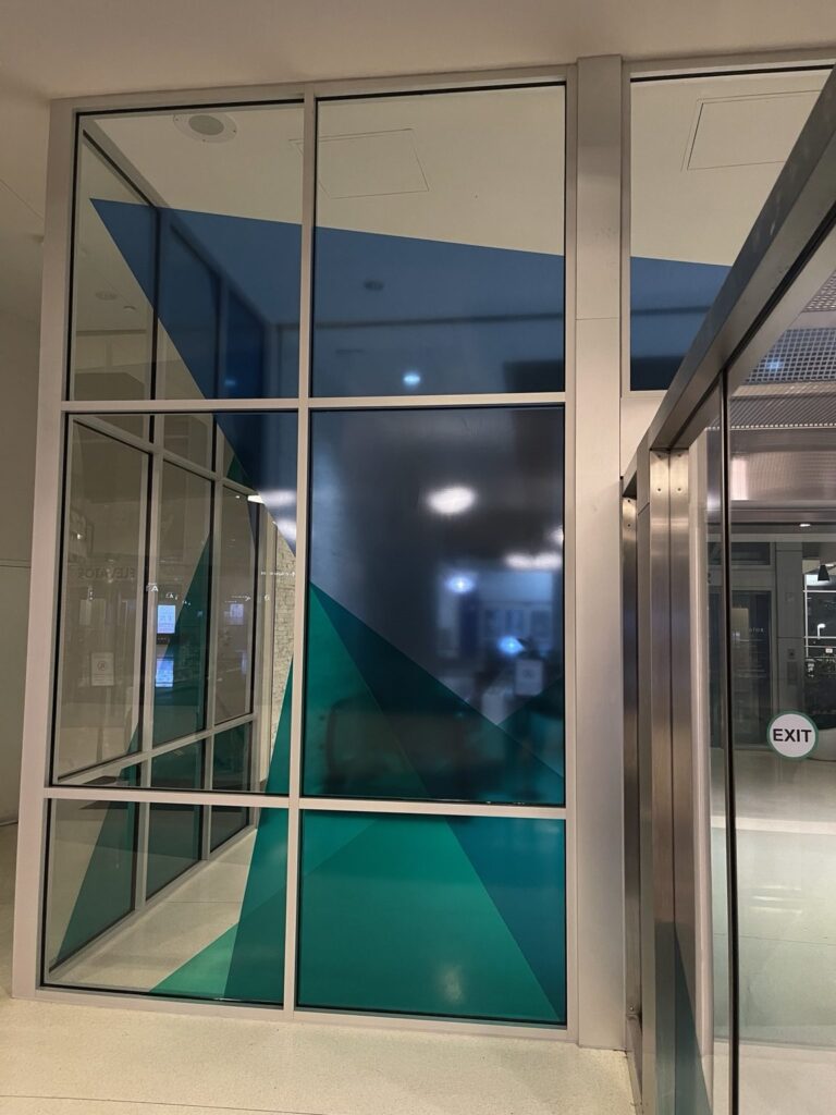 Floor-to-ceiling airport glass wall with dark blue and teal geometric vinyl shapes aligned across multiple aluminum-framed window panels.