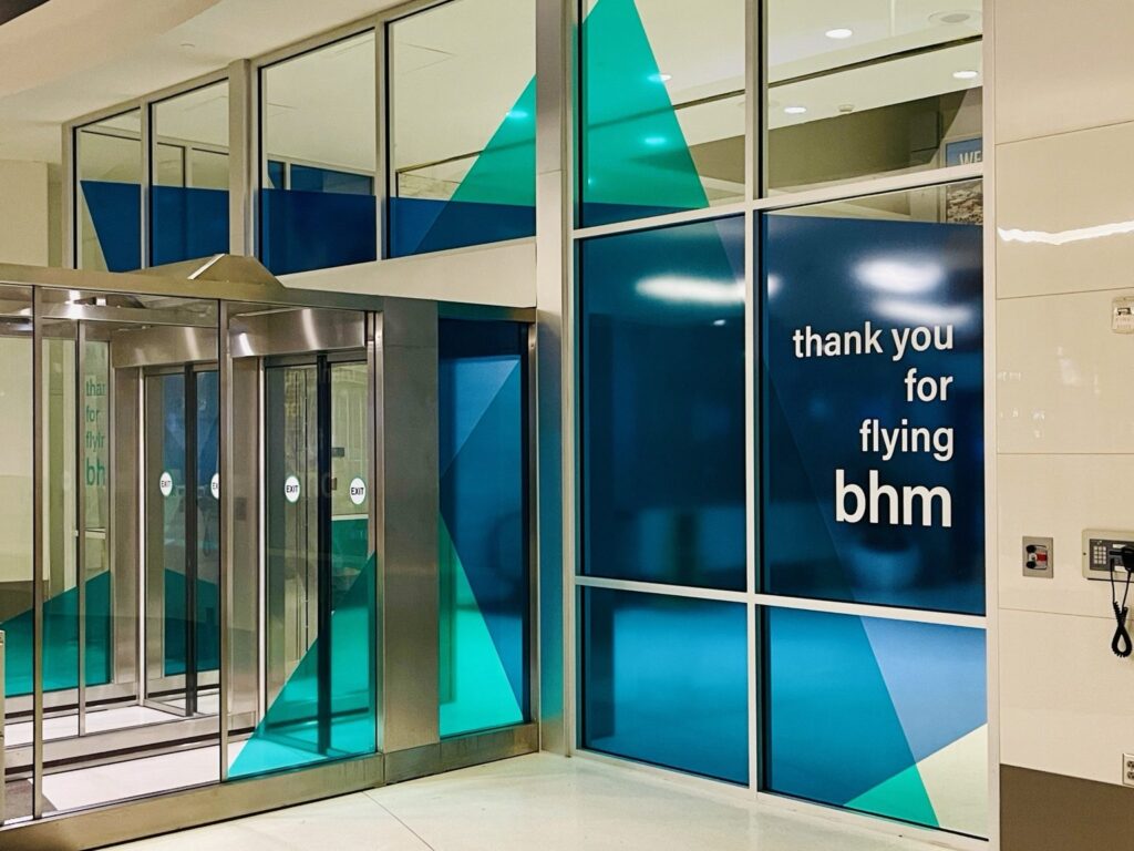 Birmingham airport window graphics: wide view of the Birmingham airport entrance with geometric teal and navy vinyl graphics spanning the nearby multi-pane glass wall.