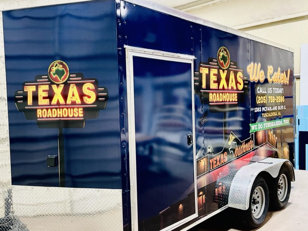 Three-quarter view of a completed Texas Roadhouse promotional cargo trailer with bold yellow and red restaurant logos on a deep blue vinyl wrap.