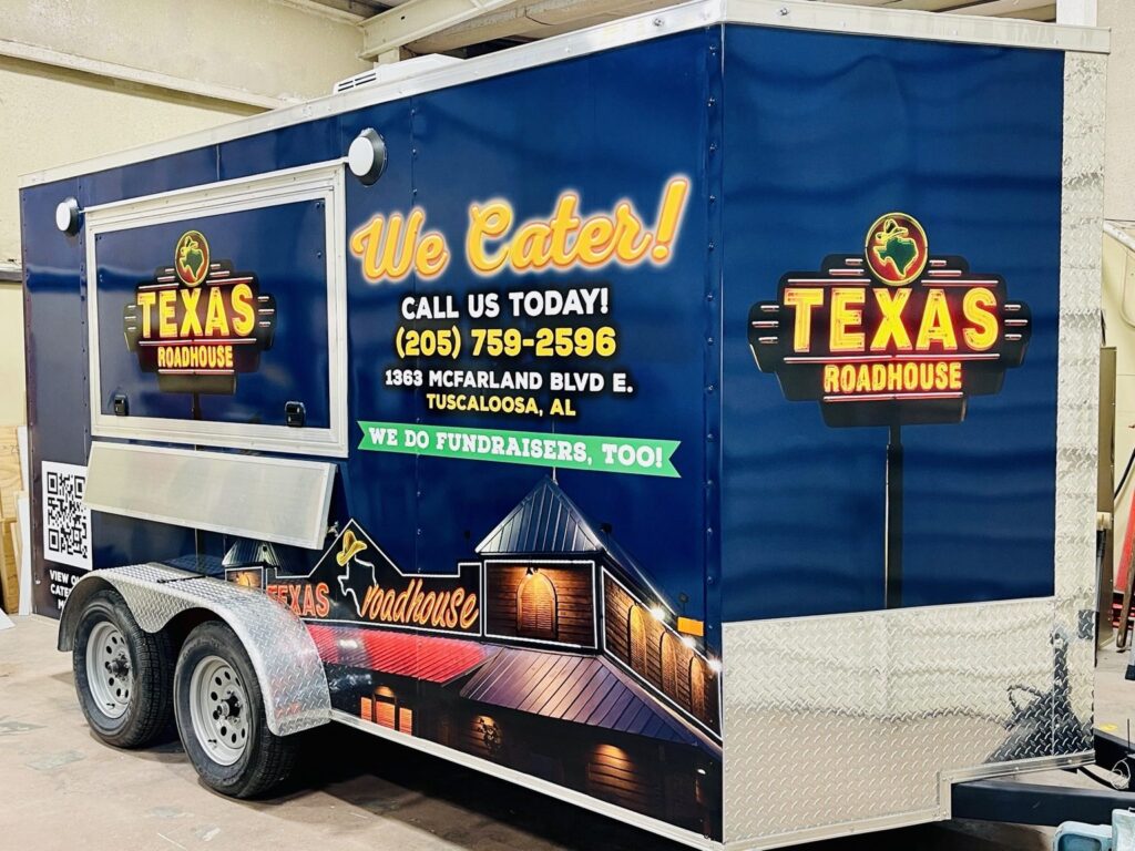 Side and front of a Texas Roadhouse catering trailer featuring custom printed restaurant branding, catering messaging, a QR code and printed building graphics.