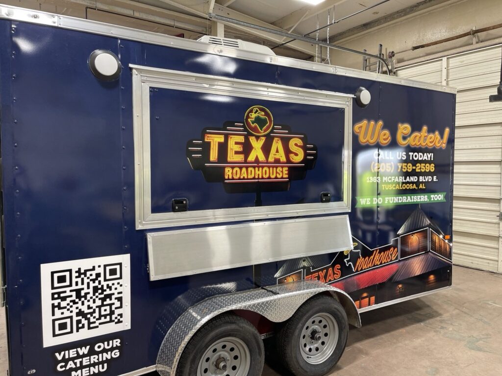 Enclosed dual-axle Texas Roadhouse trailer showing full-color navy 3M IJ180 wrap graphics around a side service window and aluminum diamond-plate fenders.