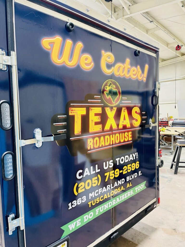Rear double doors of a Texas Roadhouse cargo trailer with a custom printed We Cater message, large Texas Roadhouse logo and fundraising graphics.