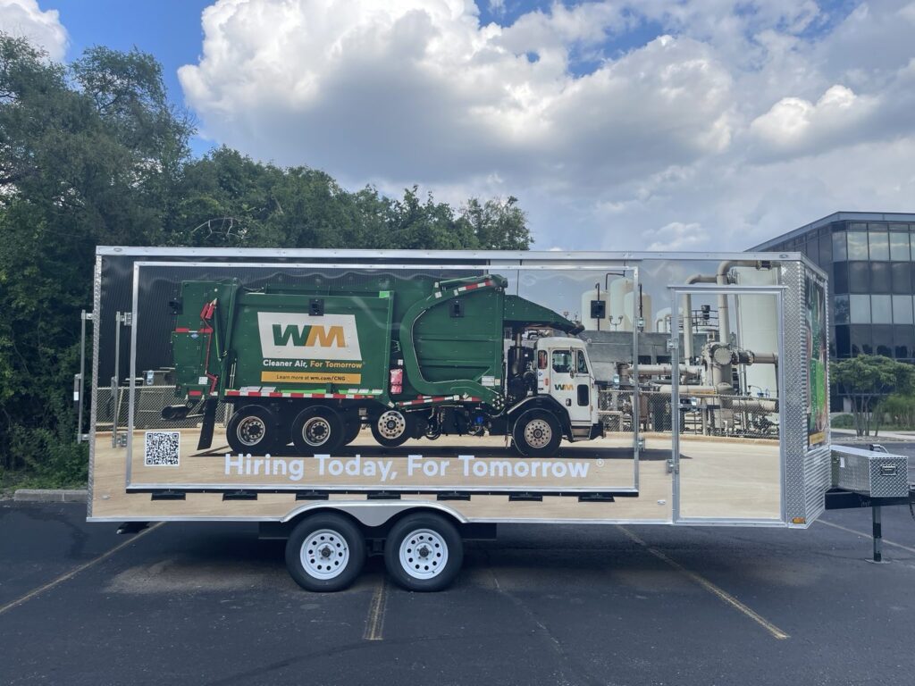 Full side profile of the WM cargo trailer with a large vehicle graphic and employment messaging