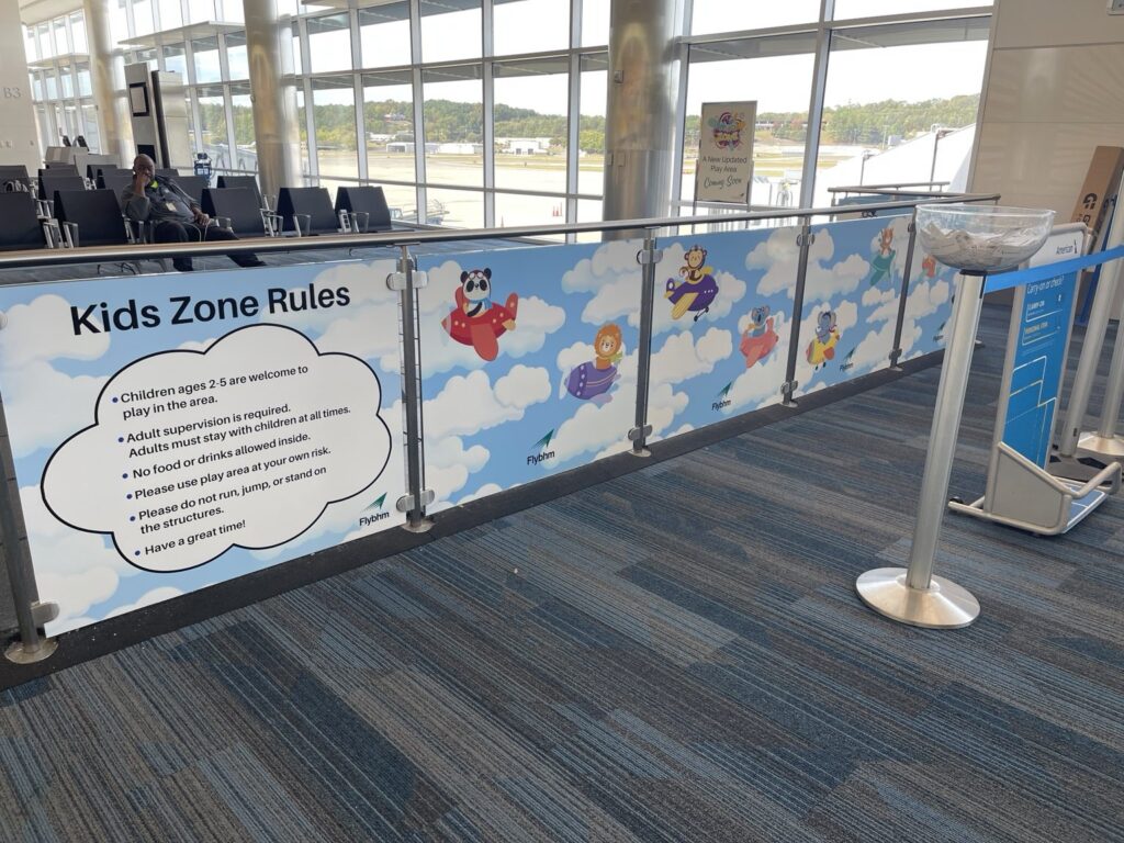 Long angled view of cartoon airplane and cloud graphics on panels around an airport Kids Zone, with passenger seating and windows beyond.