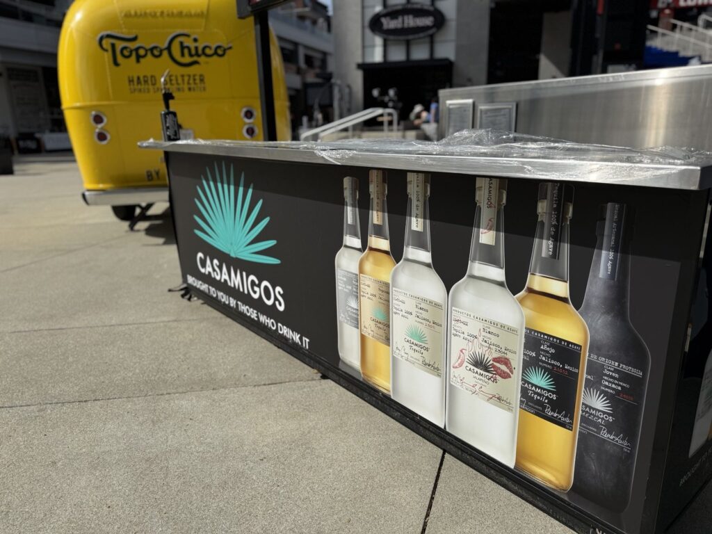 Branded Casamigos stadium counter wrap with turquoise agave icon, white Casamigos lettering, and printed bottle lineup on a black background.