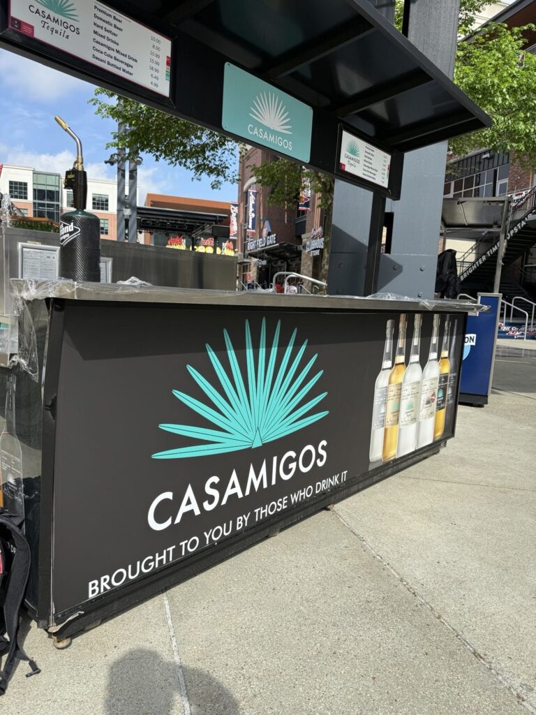 Angled front view of black Casamigos concession cart featuring a large turquoise agave symbol, white lettering and printed bottle images beneath menu boards.