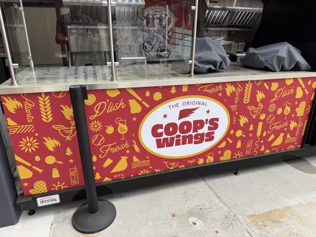 Front-facing view of Coop's Wings concession counter with red and yellow patterned wrap, white central logo, and glass food-service barrier.