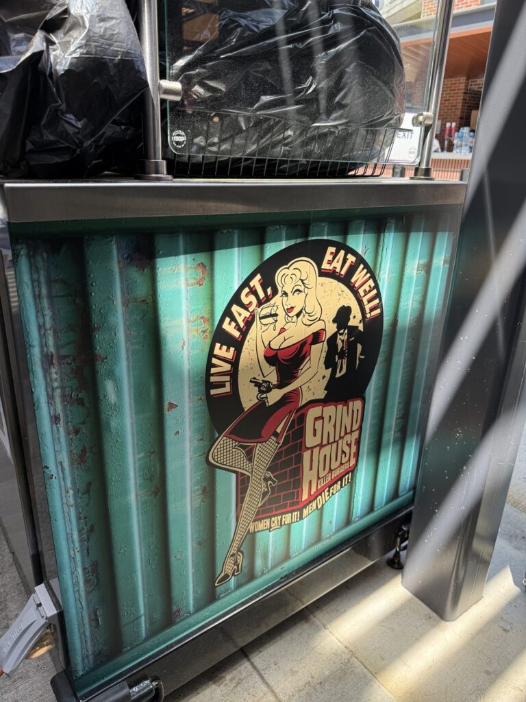 Close-up of Grindhouse Killer Burgers concession cart graphic showing vintage illustrated character, Live Fast Eat Well slogan, and distressed teal background.