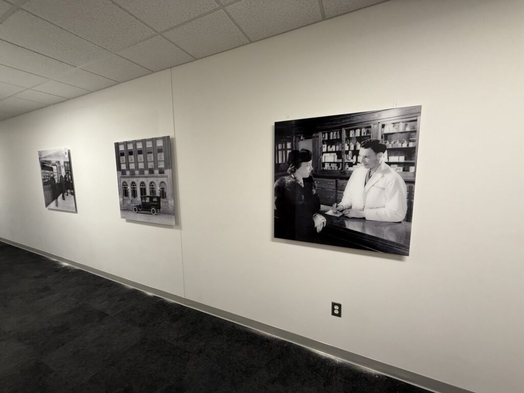 Three large black-and-white photographic panels installed along a white hallway wall, including an old pharmacy scene and historic building.