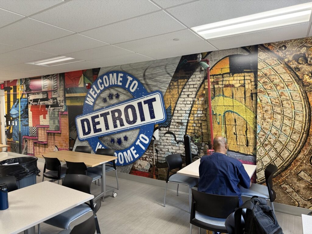 Detroit wall wrap: commercial break room featuring a colorful full-wall Welcome to Detroit mural with urban brick and graffiti imagery behind tables and chairs.