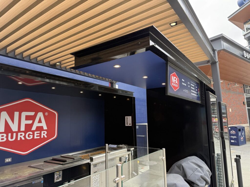 Angled view of NFA Burger concession area with dark blue wall, red-and-white NFA Burger badge, black kiosk structure and menu board.