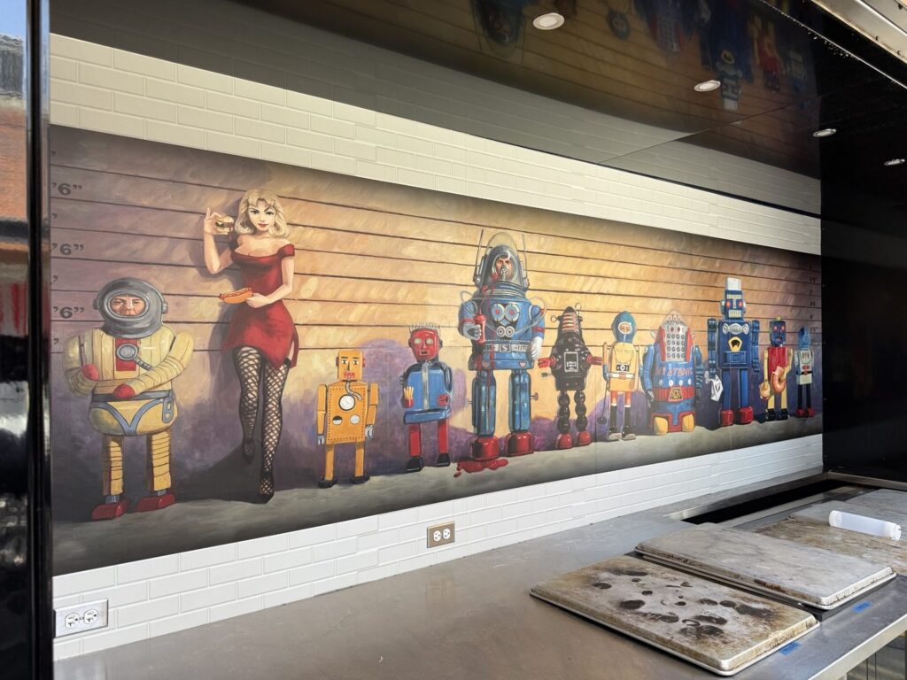 Wide interior view of illustrated concession wall graphic showing vintage robots, retro characters, and a pin-up style figure above stainless steel counters.