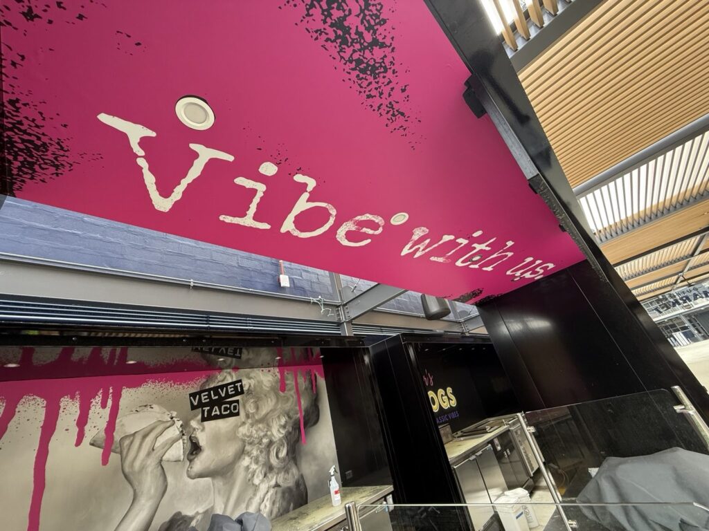 Upward view of bright magenta Velvet Taco overhead graphic with large Vibe with us lettering and black distressed details.